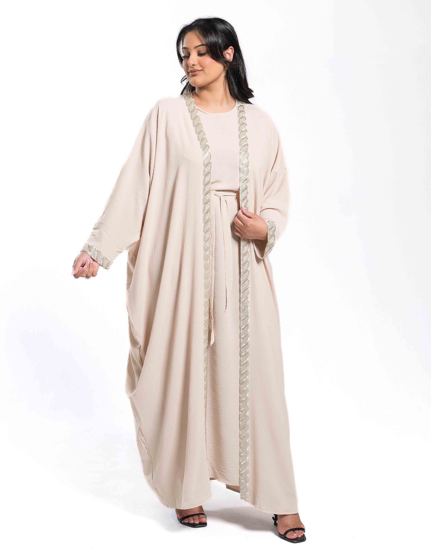 A woman wearing a beige batwing abaya showing the full front view with flowing fabric and design details.