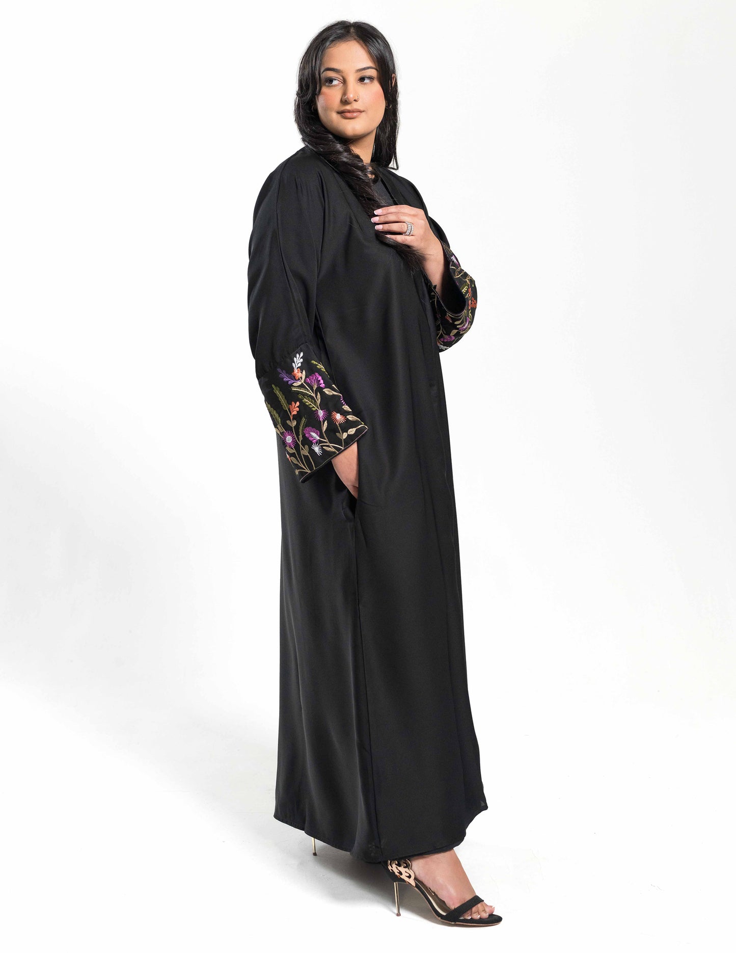 A woman wearing a black floral bloom abaya with hand-in-pocket pose, featuring detailed embroidery and a matching belt.