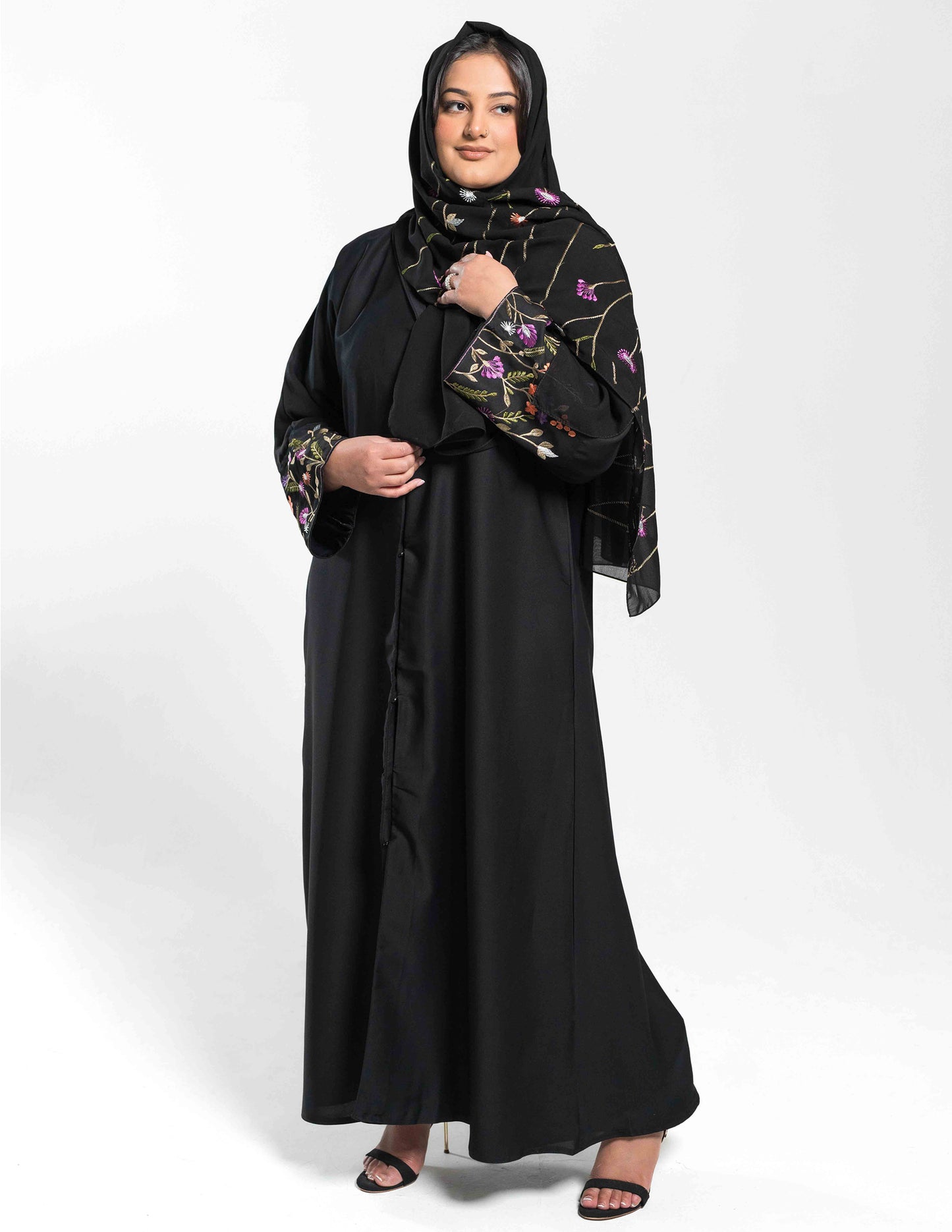 A woman wearing a black abaya with blazer like lapels and floral hand-embroidery on the sleeves, paired with a matching belt and hijab.