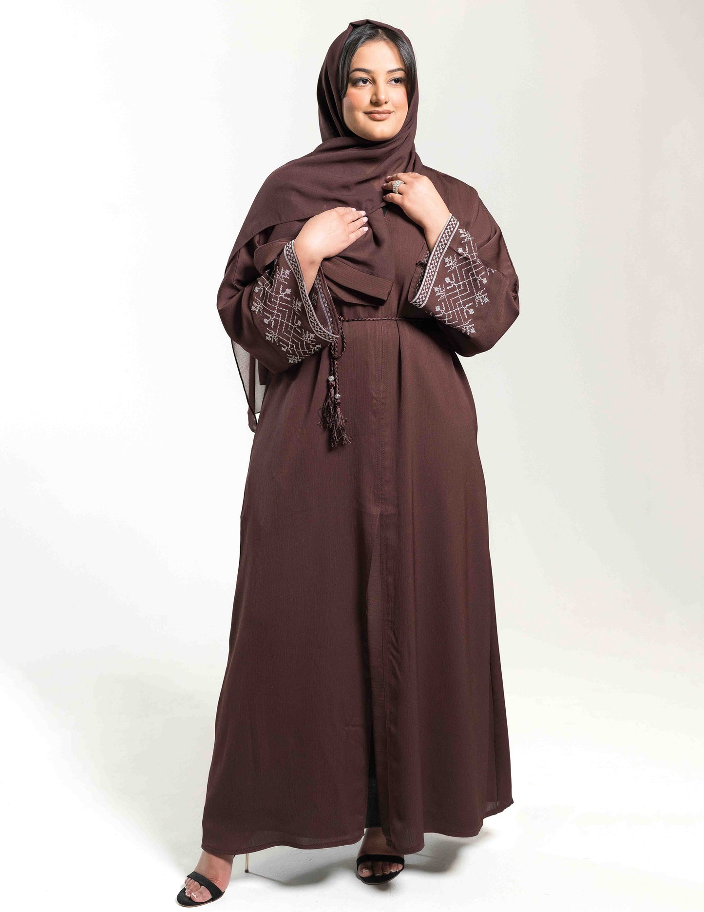 A woman wearing a brown open abaya with white embroidery on the sleeves, paired with a matching hijab and belt.