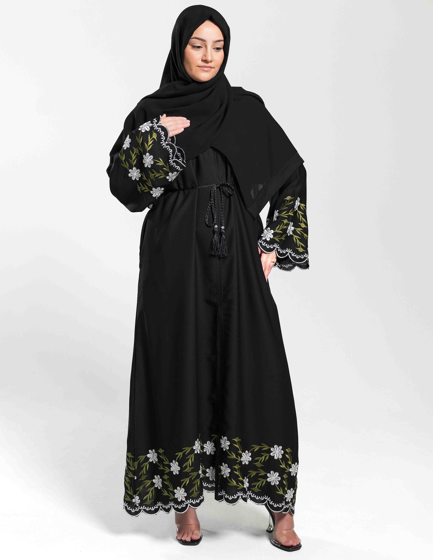 A woman wearing a black open abaya with green and white hand-embroidery, including floral patterns along the sleeves and bottom border.