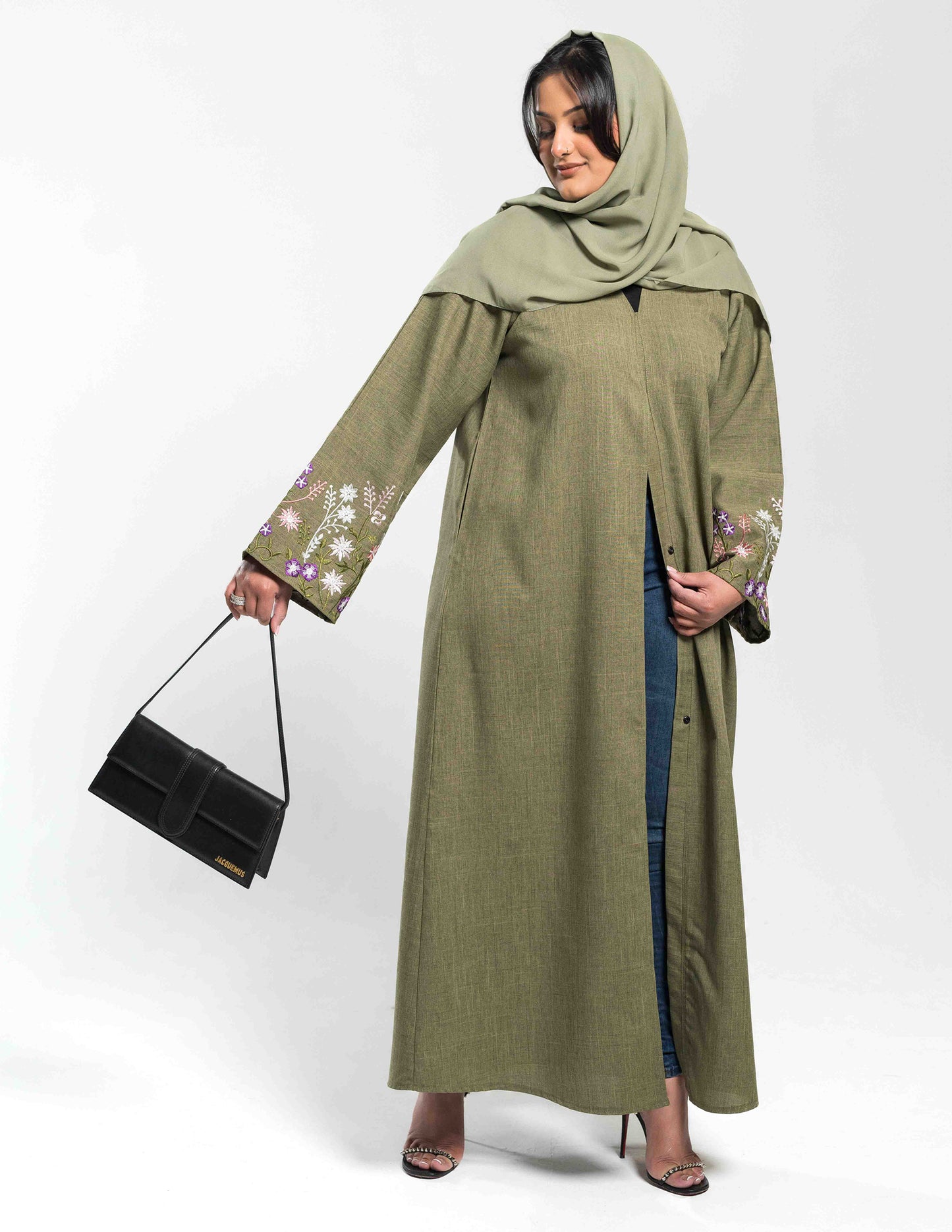 A woman wearing a green linen Zahra abaya holding a bag, showing the front view with flowing fabric and design details.
