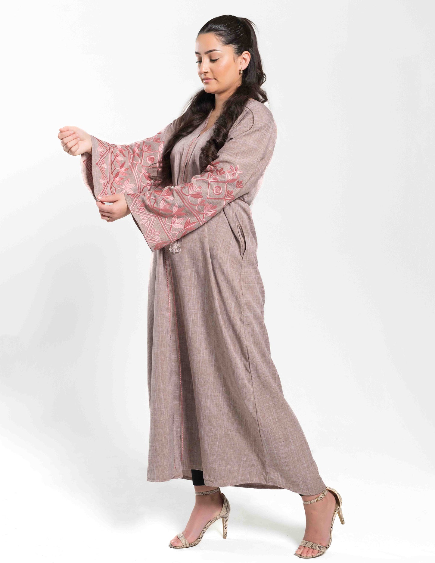 A woman wearing a beige abaya with pink embroidery on the sleeves and a matching hijab and belt.