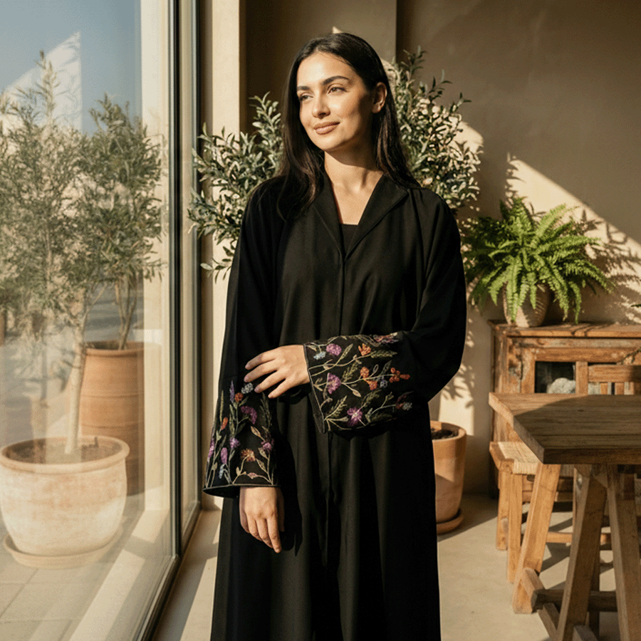 Woman in a black outfit with floral embroidery standing in a sunlit room with plants.