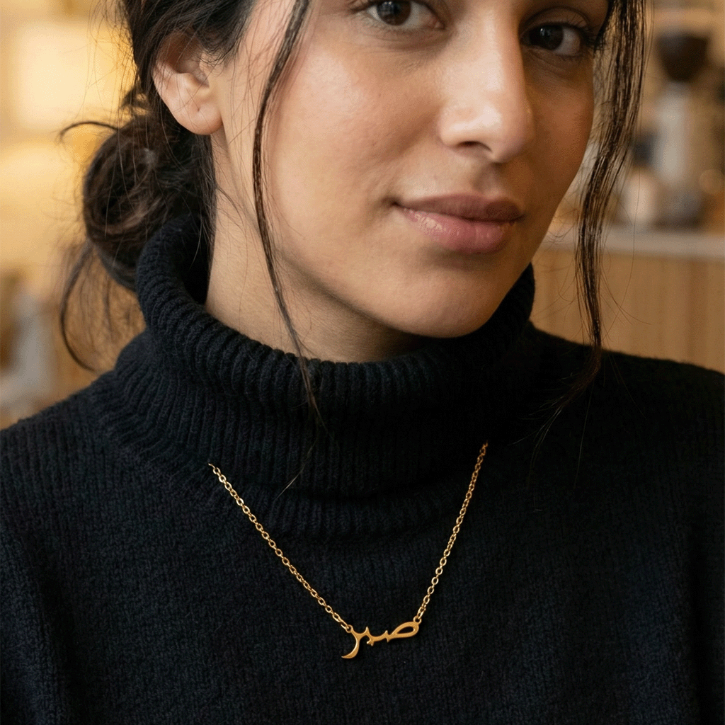 Woman wearing a black turtleneck and gold sabr necklace with Arabic script.
